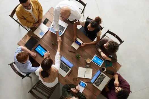 Overhead view of diverse team collaborating with laptops and tablets around a wooden conference table in a modern office, focusing on teamwork and digital communication.