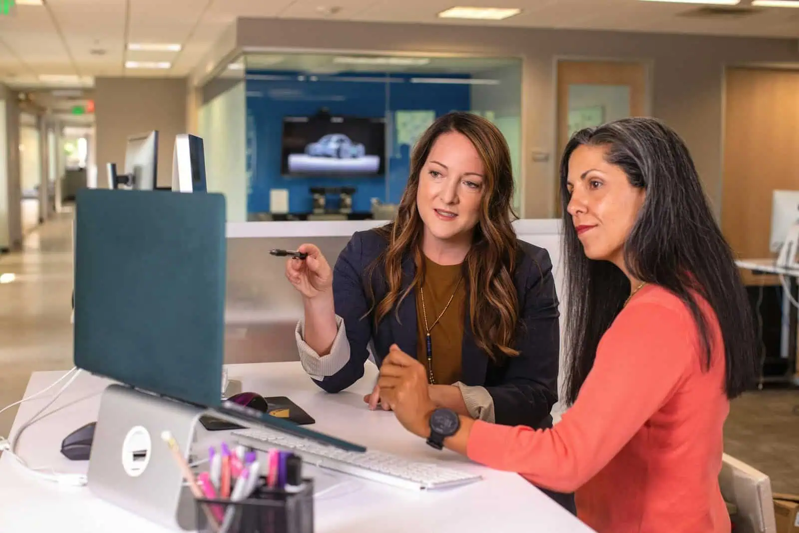 Professional women discussing work at a desk in an office setting, collaborating on a project, using a computer, representing teamwork and productivity in the workplace.