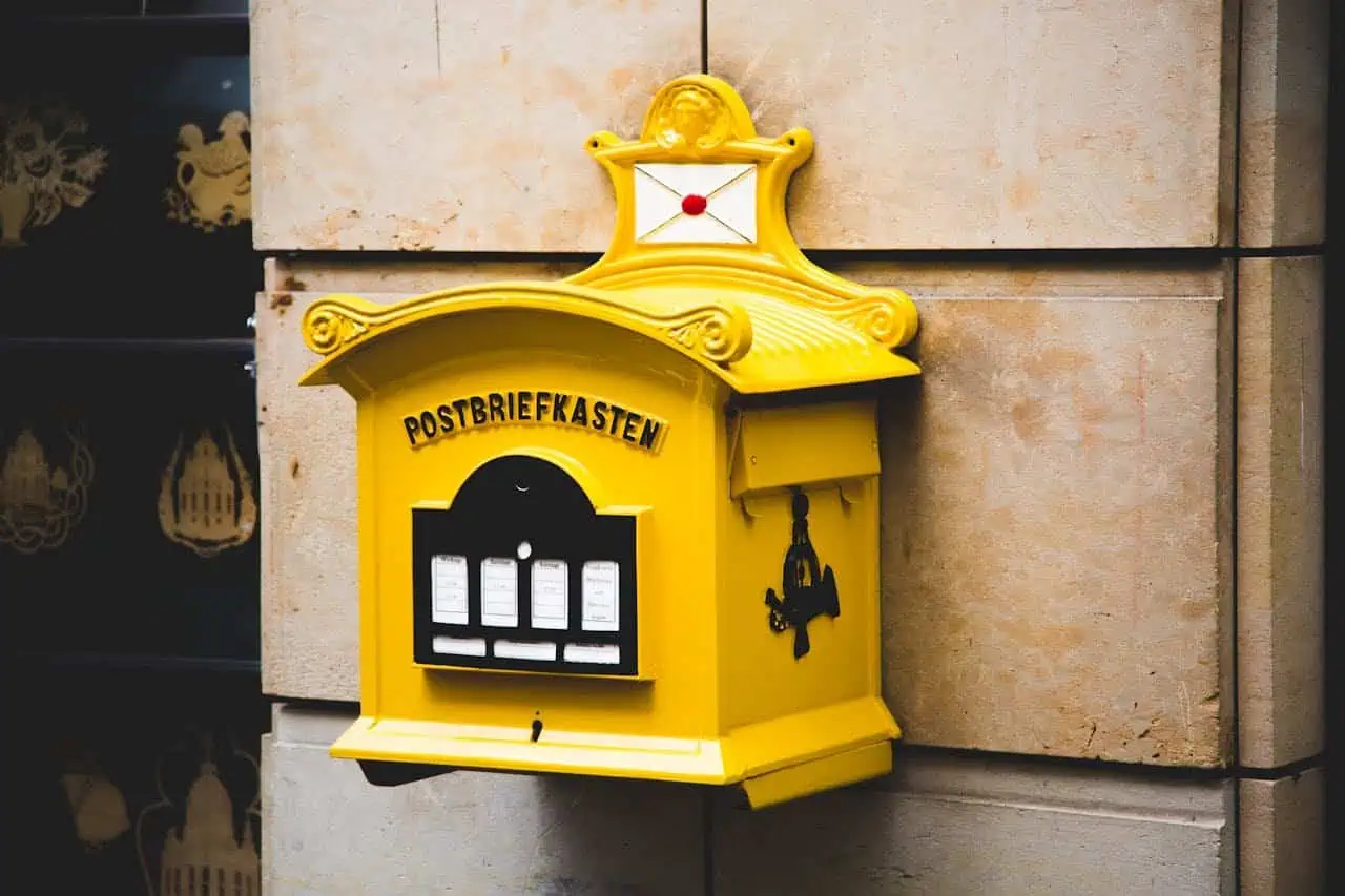Yellow postbox with black and white display for mail collection, mounted on beige stone wall in an urban setting.