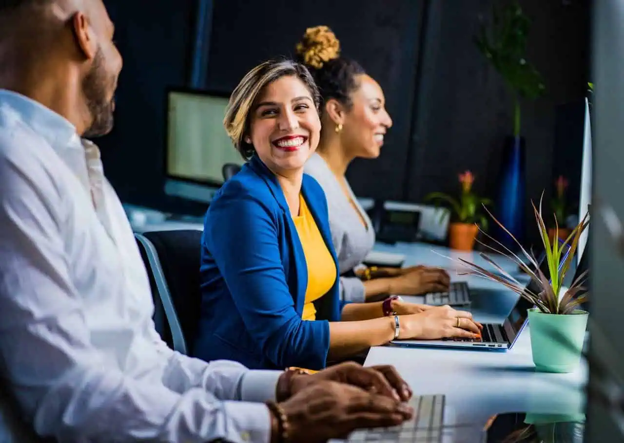 Smiling diverse business team working together at a modern office desk with laptops and plants.
