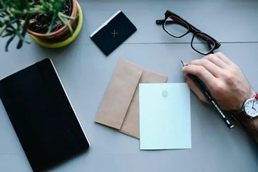 A person writing on a desk with notebooks, greeting cards, eyeglasses, and a potted plant, showcasing a workspace setup for creativity or planning.