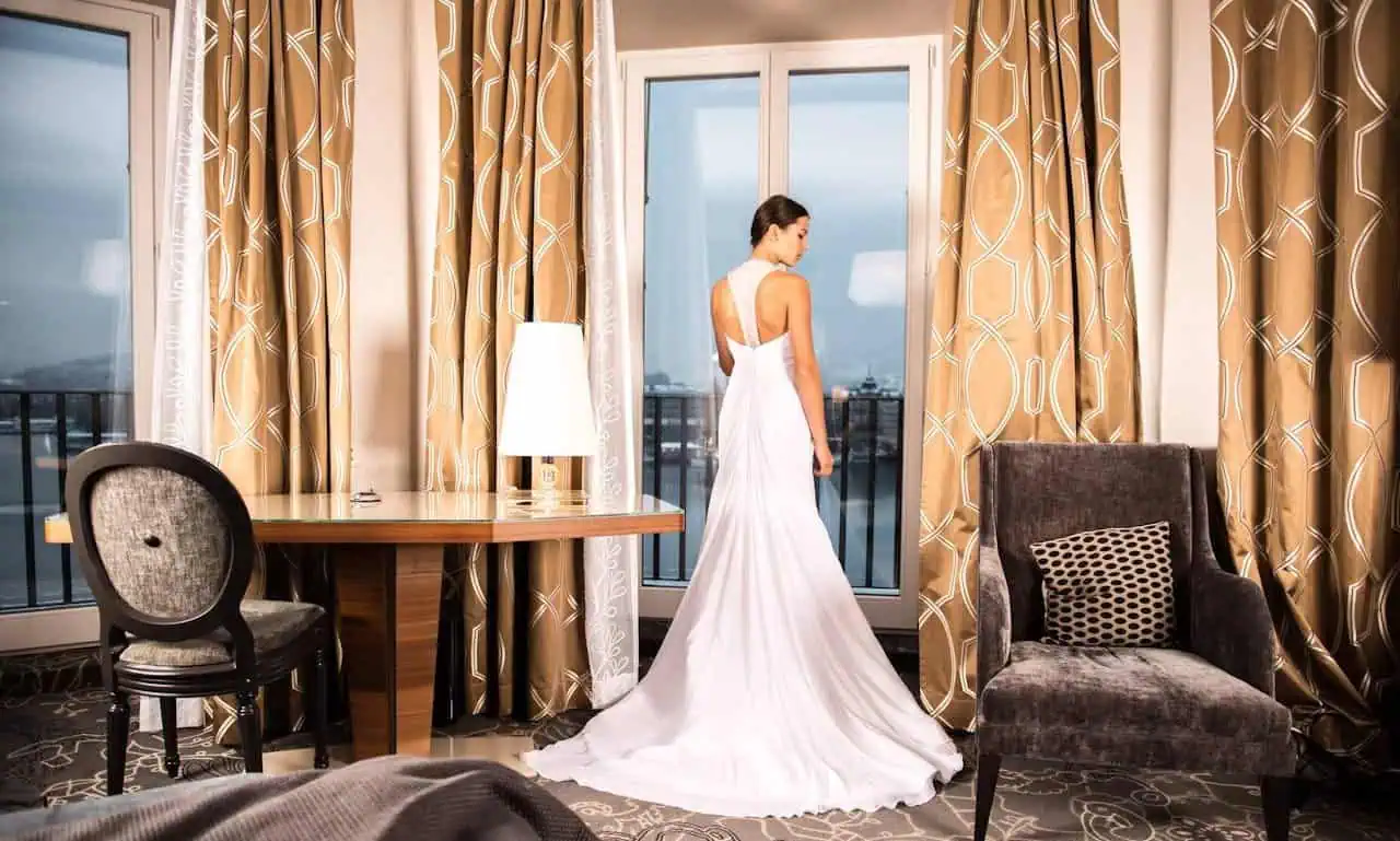 Elegant bride in a white wedding gown standing by a large glass door in a stylish hotel room with gold curtains and modern furniture.