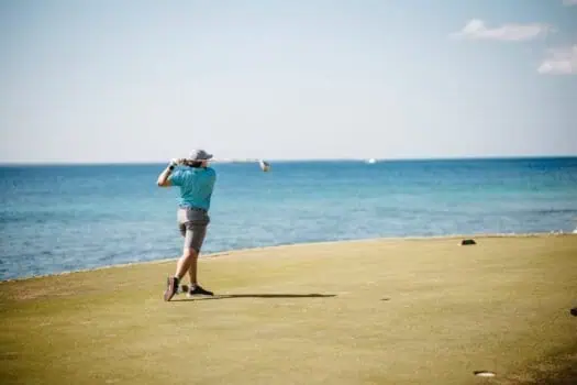 Golf player hitting a shot on a seaside golf course with ocean view, sunny sky, and clear weather, illustrating leisure, golf, and coastal outdoor activities.