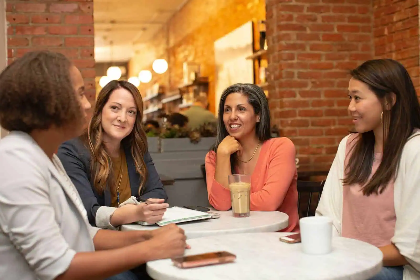 Friendly women’s support group discussion in cozy coffee shop, diverse community, mental health awareness, peer support, social connection, empowering conversations.