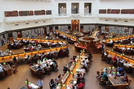 Brightly lit library with many people studying at wooden desks, surrounded by tall bookshelves, showcasing a historic and spacious reading environment.