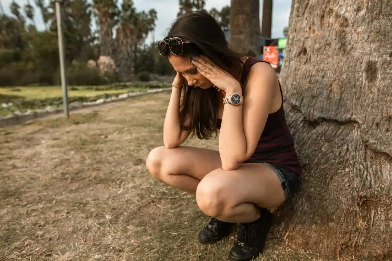 Distressed woman sitting on ground by tree, holding head in hands, expressing worry or stress, outdoor park setting with grass and trees, emotional moment of distress or anxiety, relevant for mental health support and wellbeing content.
