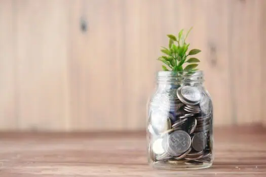 Growing plant emerging from a jar filled with coins, symbolising financial growth and savings, on a wooden background.