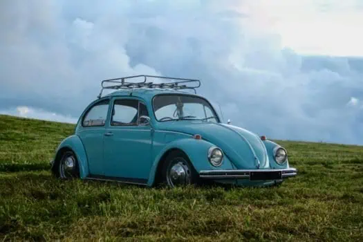 Blue vintage Volkswagen Beetle with a roof rack parked on a grassy field under a cloudy sky.