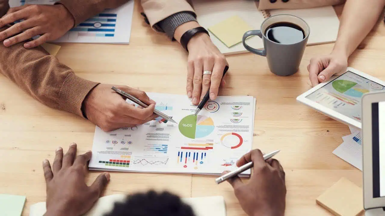 High-angle view of business professionals analysing graphs and data charts during a team meeting at a wooden table, with a coffee mug and digital tablet nearby, promoting teamwork and data analysis expertise.