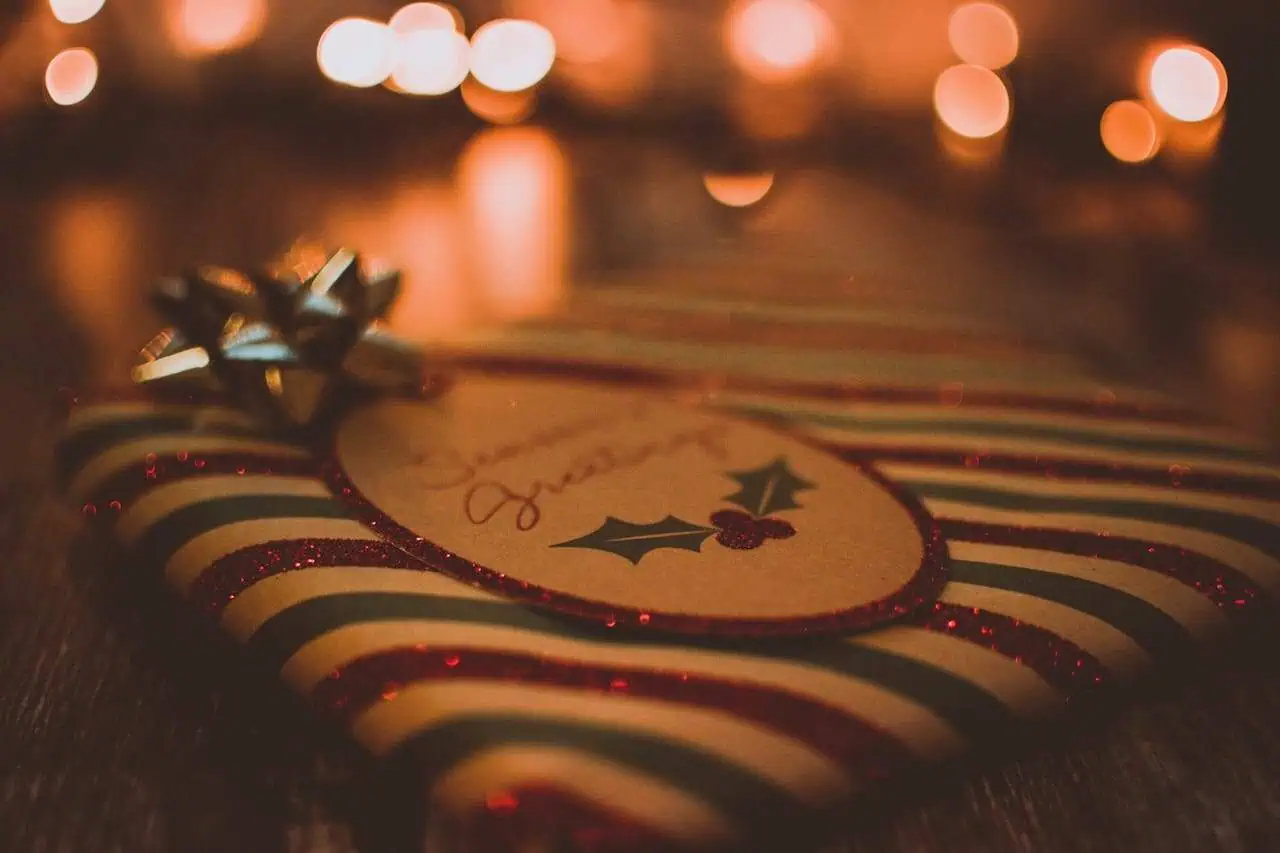 Glittery festive Christmas ornament with holly design and handwritten message, blurred warm background with bokeh lights, holiday decoration, seasonal celebration, festive decor, Prowess UK.