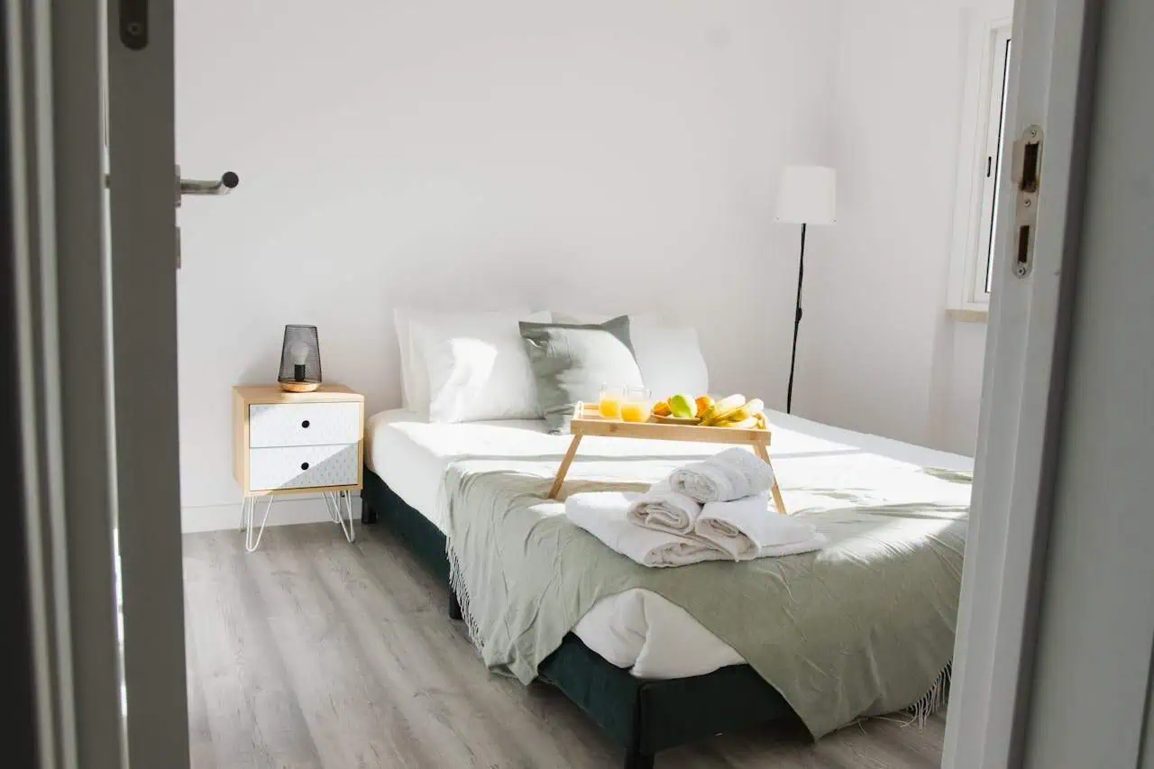 Bright minimalist bedroom with white walls, neatly made bed with fresh towels, natural light, and modern decor, ideal for a comfortable stay.