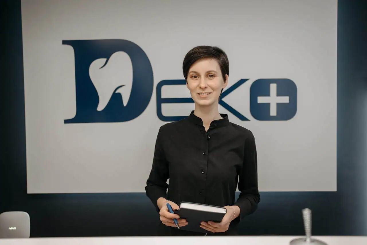 Smiling young professional woman standing in front of Prowess Dental Clinic logo, holding a notepad and pen, representing dental care, healthcare professionalism, and patient service.