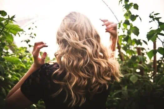 Blonde woman with curly hair outdoors in a lush green garden or greenhouse, enjoying nature and sunlight interaction, showing a connection with environment and well-being.