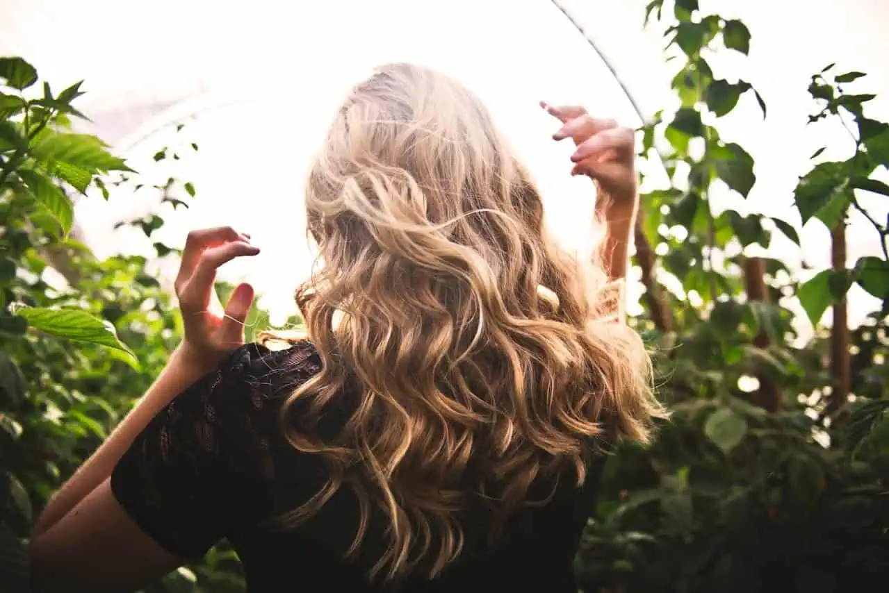 Blonde woman with curly hair outdoors in a lush green garden or greenhouse, enjoying nature and sunlight interaction, showing a connection with environment and well-being.