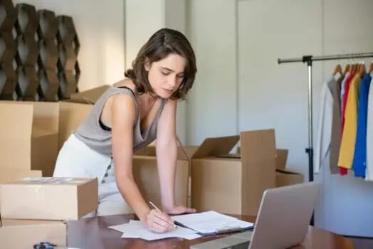 Flexible working woman sorting clothing inventory with laptop and paperwork in clothing store or warehouse environment.