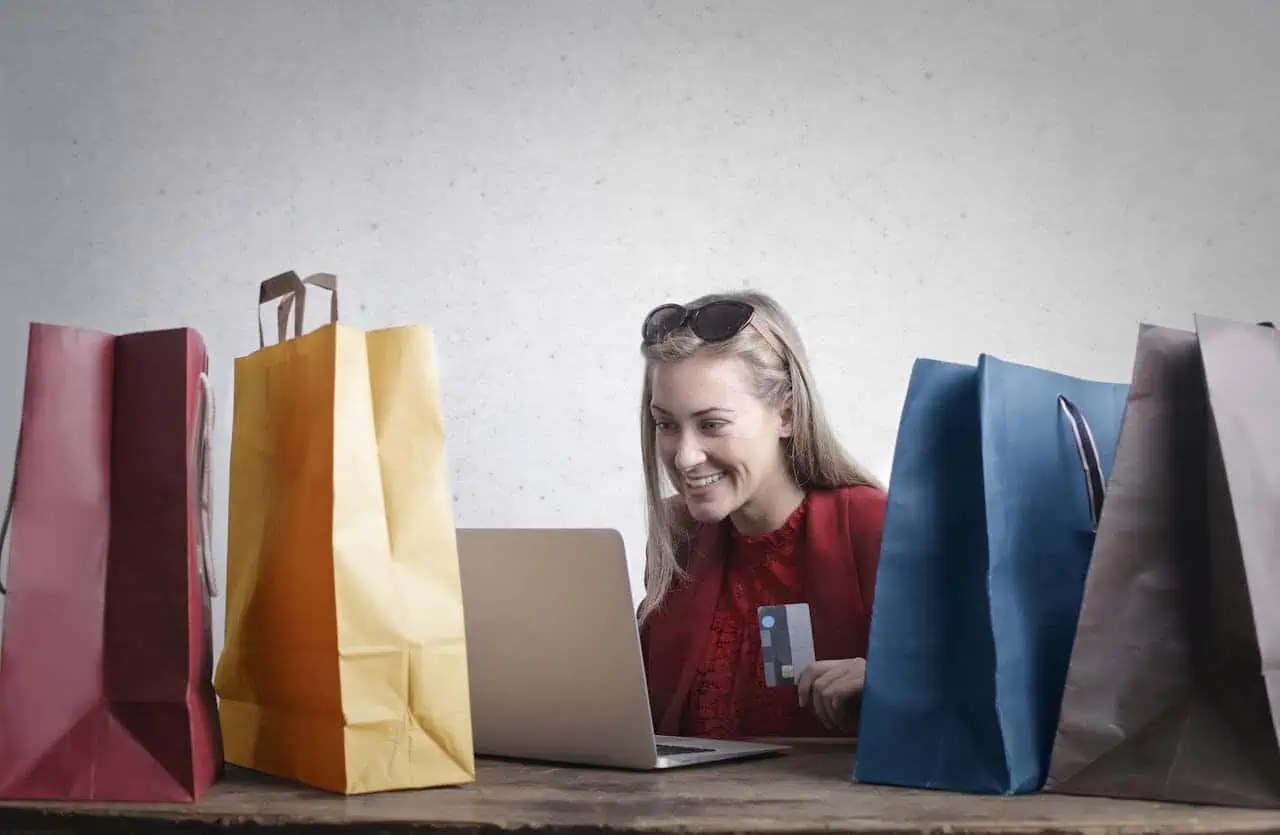 Colorful shopping bags surrounding a woman using a laptop and holding a credit card, representing online shopping, e-commerce, and digital consumer behaviour.
