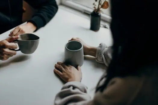 Warm beverage exchange at a modern, minimalist café table for socialising and coffee culture. A woman and a man share a moment over coffee mugs in a cosy, contemporary setting.