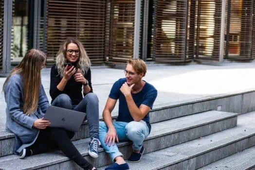 Smiling group of young people sitting on outdoor steps using laptop and mobile phone, diverse youth socialising, modern urban environment, friendship and collaboration, technology and digital communication, Prowess community event.