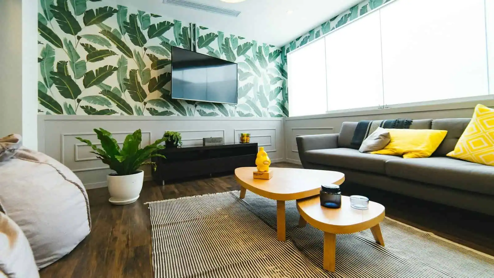 Vibrant living room with botanical wall design, modern grey sofa with yellow and neutral cushions, wooden coffee tables, large window letting in natural light, and decorative plants enhancing the space.