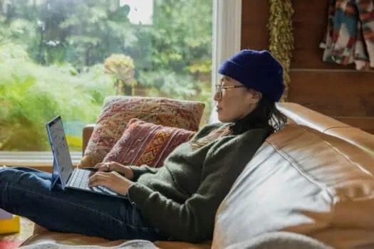 Woman using a laptop on a cosy sofa in a rustic home with large window and greenery outside, illustrating remote work and home comfort, aligning with mental health and wellbeing themes.