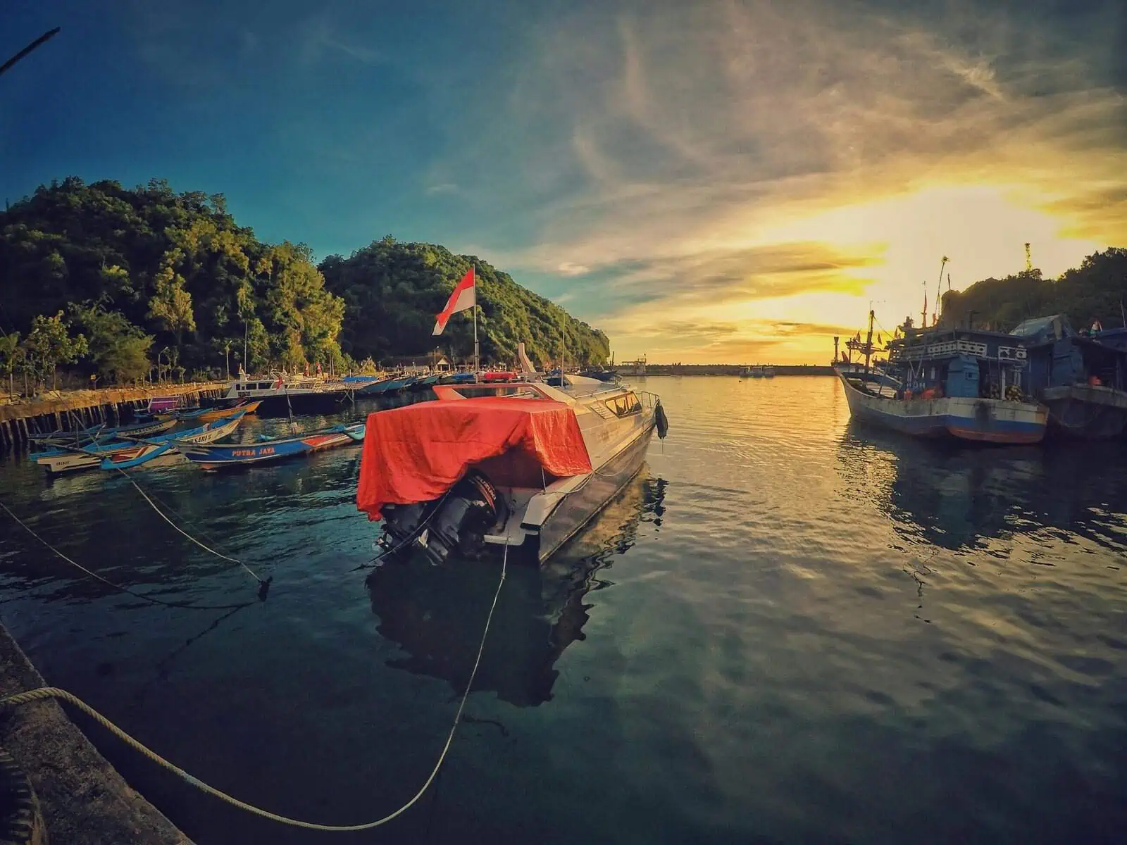 Boats docked at a serene harbour during sunset, surrounded by lush green mountains, creating a peaceful coastal scene.