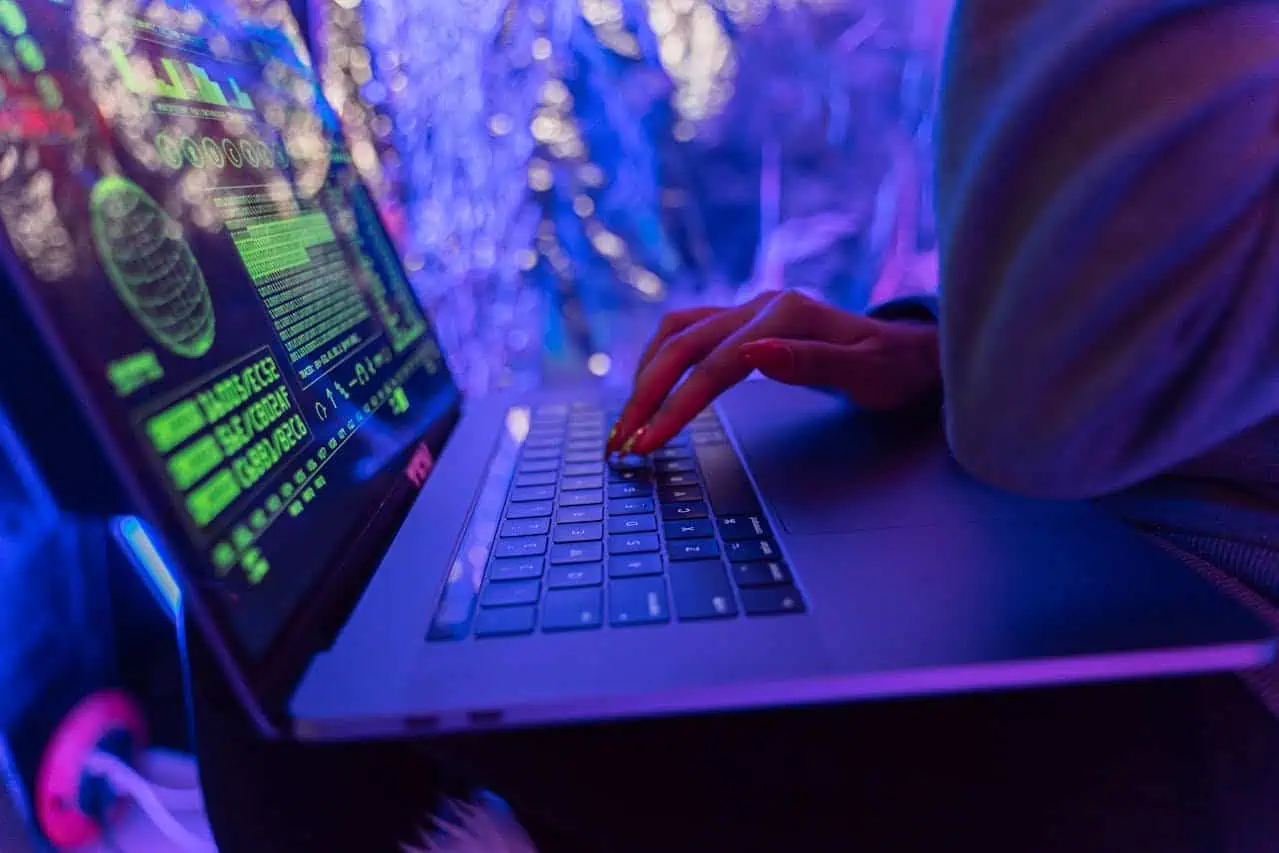 Hacker working on a laptop with cyber security code on screen in a dark, neon-lit environment.