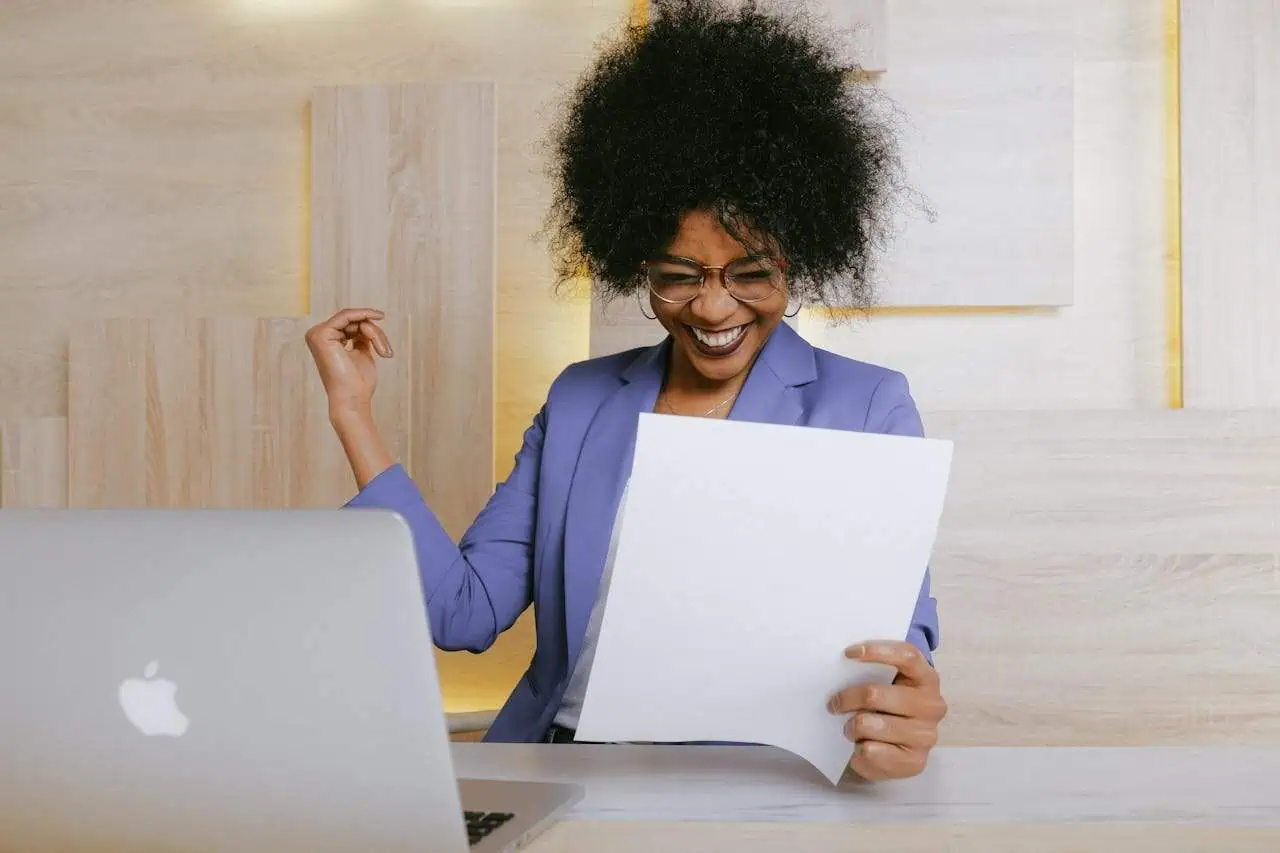 Happy woman in a business suit celebrating success while holding paper and working at her desk with a laptop.