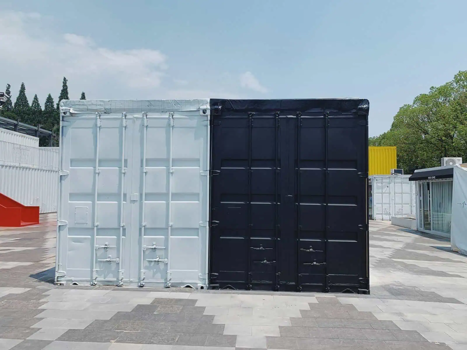 Colorful shipping containers used for storage or transport at an outdoor facility. The image features white, black, and yellow containers on a paved area with a background of trees and a blue sky.