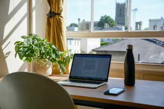 Bright home workspace featuring a laptop, a potted Monstera plant, a water bottle, and a smartphone on a wooden desk by a large window with yellow curtains, overlooking a neighbourhood with historical buildings.