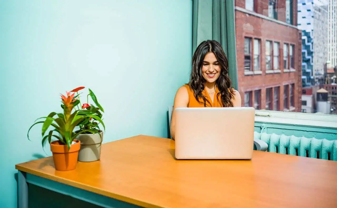 Young woman working remotely on a laptop at a sunny indoor desk with potted plants, cityscape views, and vibrant decor, promoting digital skills and career development.