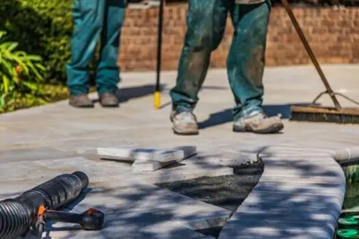 People laying paving stones on a garden pathway, with tools and construction equipment, in a residential outdoor setting.