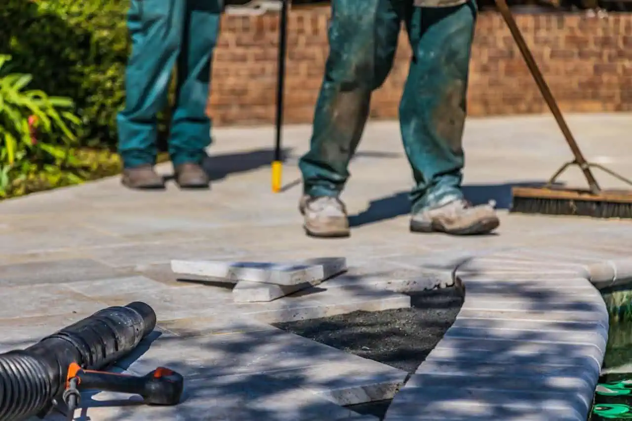 People laying paving stones on a garden pathway, with tools and construction equipment, in a residential outdoor setting.