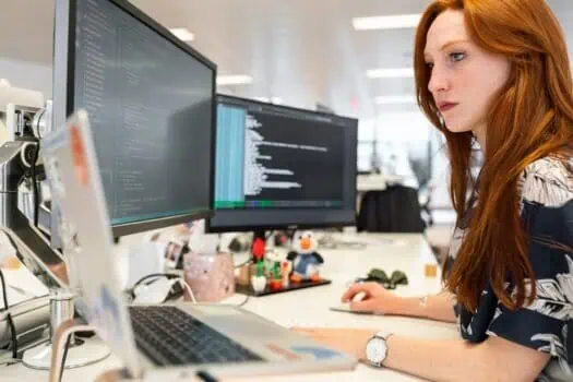 Proactive woman working on programming code at modern office desk, computer screens with coding language, female software developer, IT professional, women in tech, workplace technology environment.