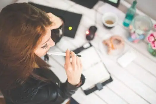 Female young woman working on creative project, brainstorming and planning with a pen in hand, surrounded by arts and crafts materials on a bright white wooden table, promoting skills development and self-empowerment.