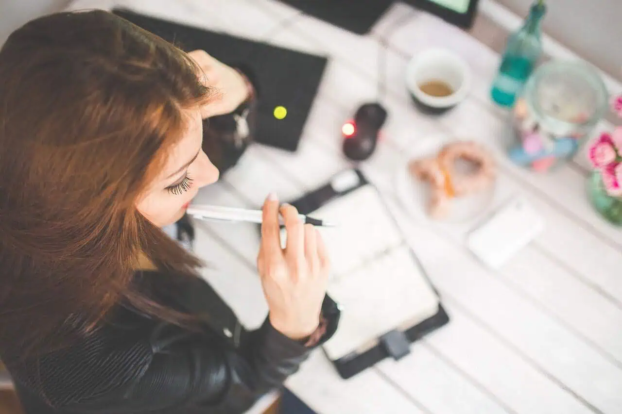 Female young woman working on creative project, brainstorming and planning with a pen in hand, surrounded by arts and crafts materials on a bright white wooden table, promoting skills development and self-empowerment.