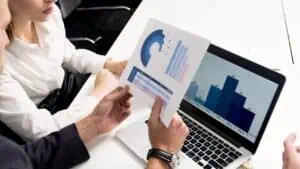 Detailed analysis of financial data and reports being reviewed by colleagues using a laptop and printed charts. Focus on data analysis, business insights, and professional collaboration.