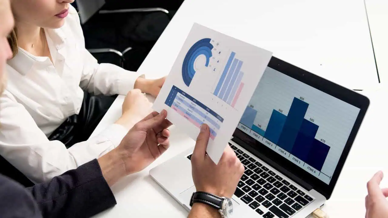 Detailed analysis of financial data and reports being reviewed by colleagues using a laptop and printed charts. Focus on data analysis, business insights, and professional collaboration.