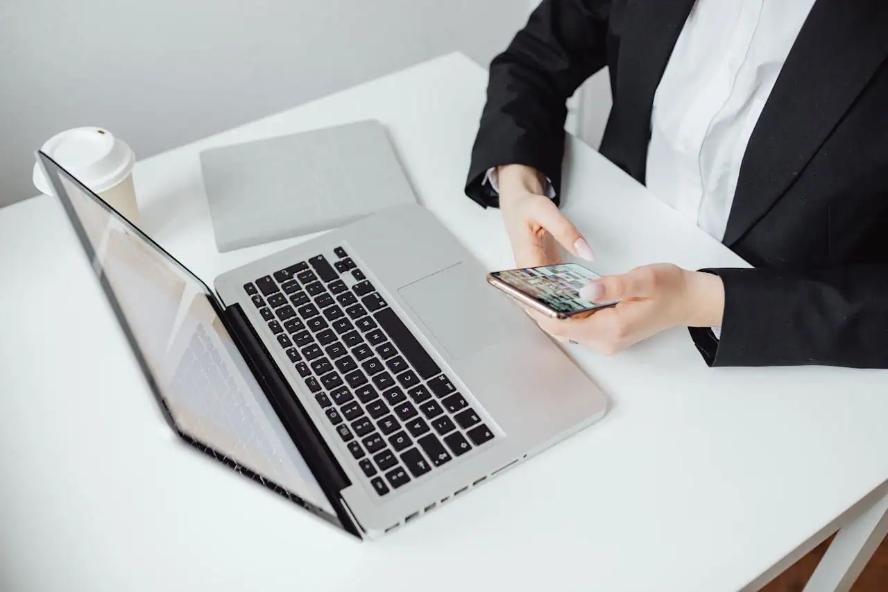 Professional woman in business suit using smartphone with open laptop and coffee cup on white desk for digital communication and remote work.
