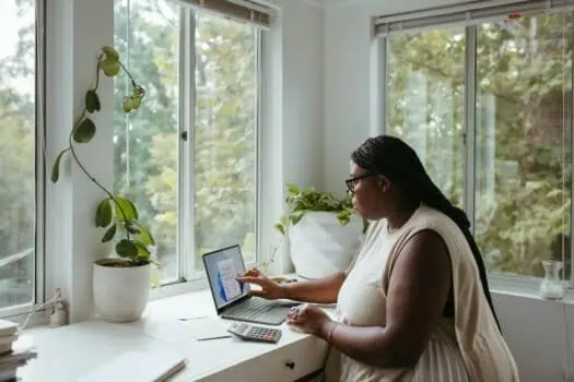 Flexible woman working on a laptop at home, studying or managing tasks in a bright room with large windows, indoor plants, and natural light, promoting remote work and productivity, concept for online learning and home office, Prowess UK.