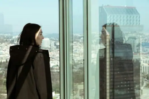 Contemplative woman looking at city skyline through glass window, reflecting urban landscape and modern architecture, symbolising introspection and connection with city life.