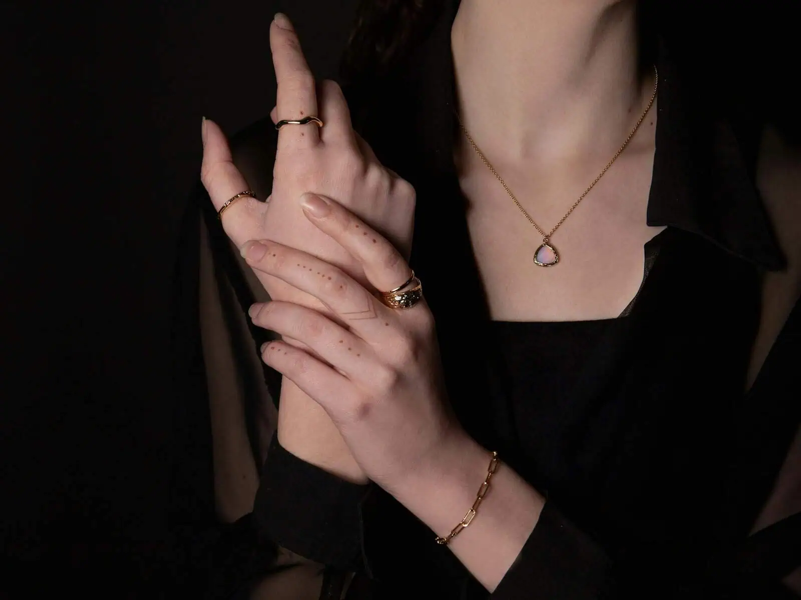 Elegant woman's hands adorned with gold rings, bracelet, and a delicate necklace featuring a heart-shaped pendant, against a dark background, showcasing stylish jewellery and fashion accessories.