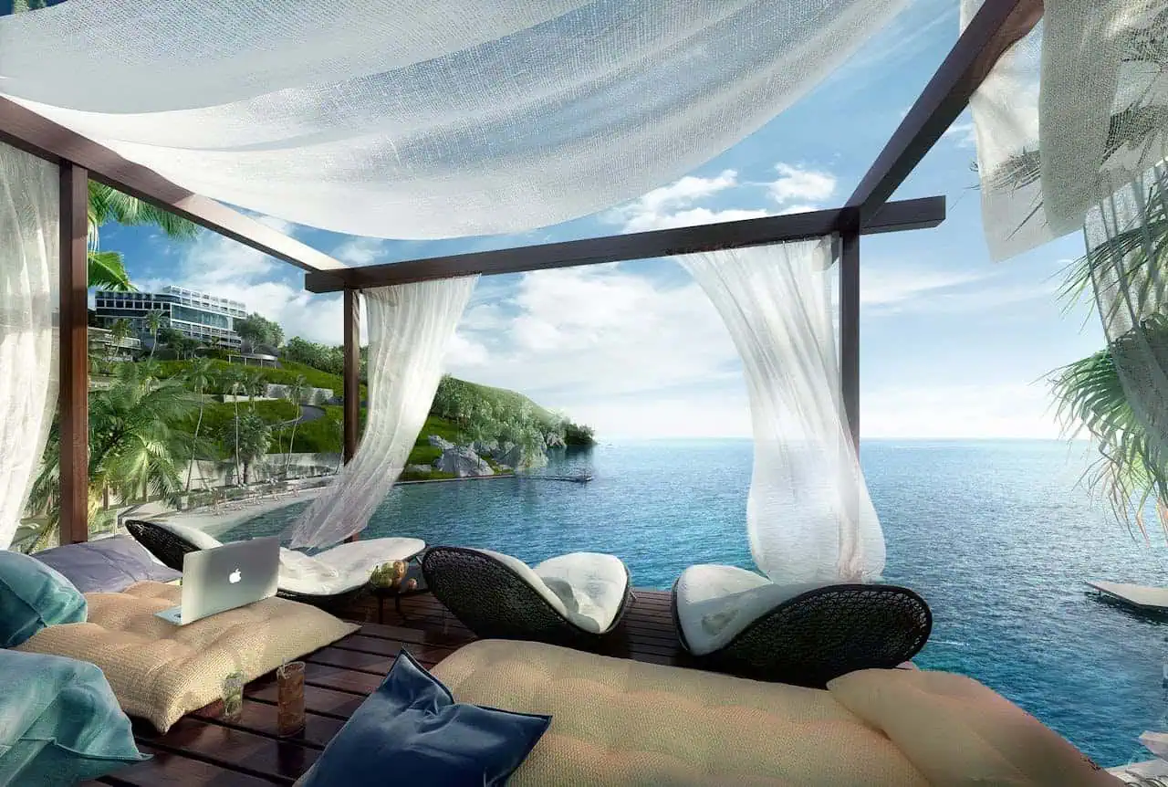 Relaxing seaside balcony with curtains, lounge chairs, and a laptop overlooking the ocean and lush hillside; perfect for tropical getaways and luxury oceanfront stays.
