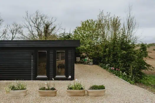 Modern black garden shed with glass doors surrounded by flower pots, trees, and a gravel patio in a peaceful outdoor space.