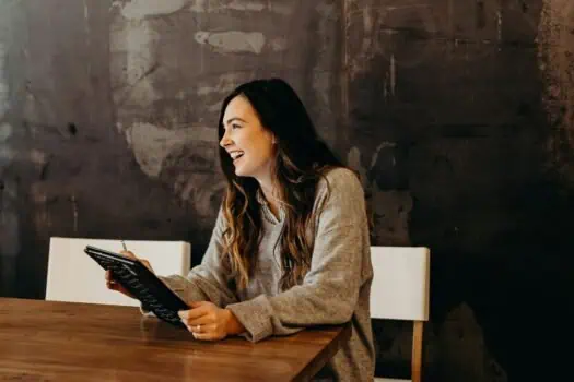 Friendly woman reading menu or tablet, seated at a wooden table in a modern, cosy café setting with warm lighting and a dark textured wall, enjoying a casual coffee break.