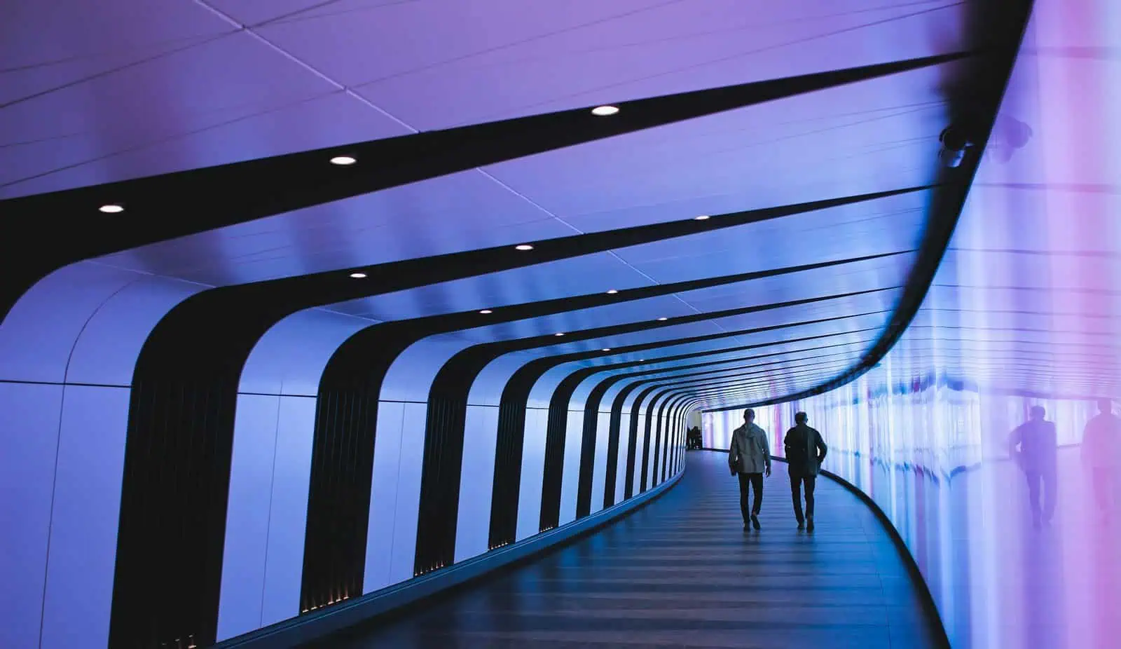 Futuristic corridor with blue lighting and two people walking, modern technology, innovative architecture, sleek design, illuminated walkway, high-tech environment, urban infrastructure, contemporary interior design, advanced construction.