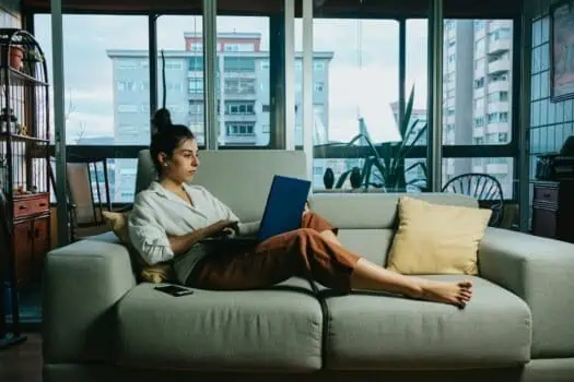 Young woman working on a laptop while relaxing on a sofa in a modern apartment, overlooking city buildings through large windows.