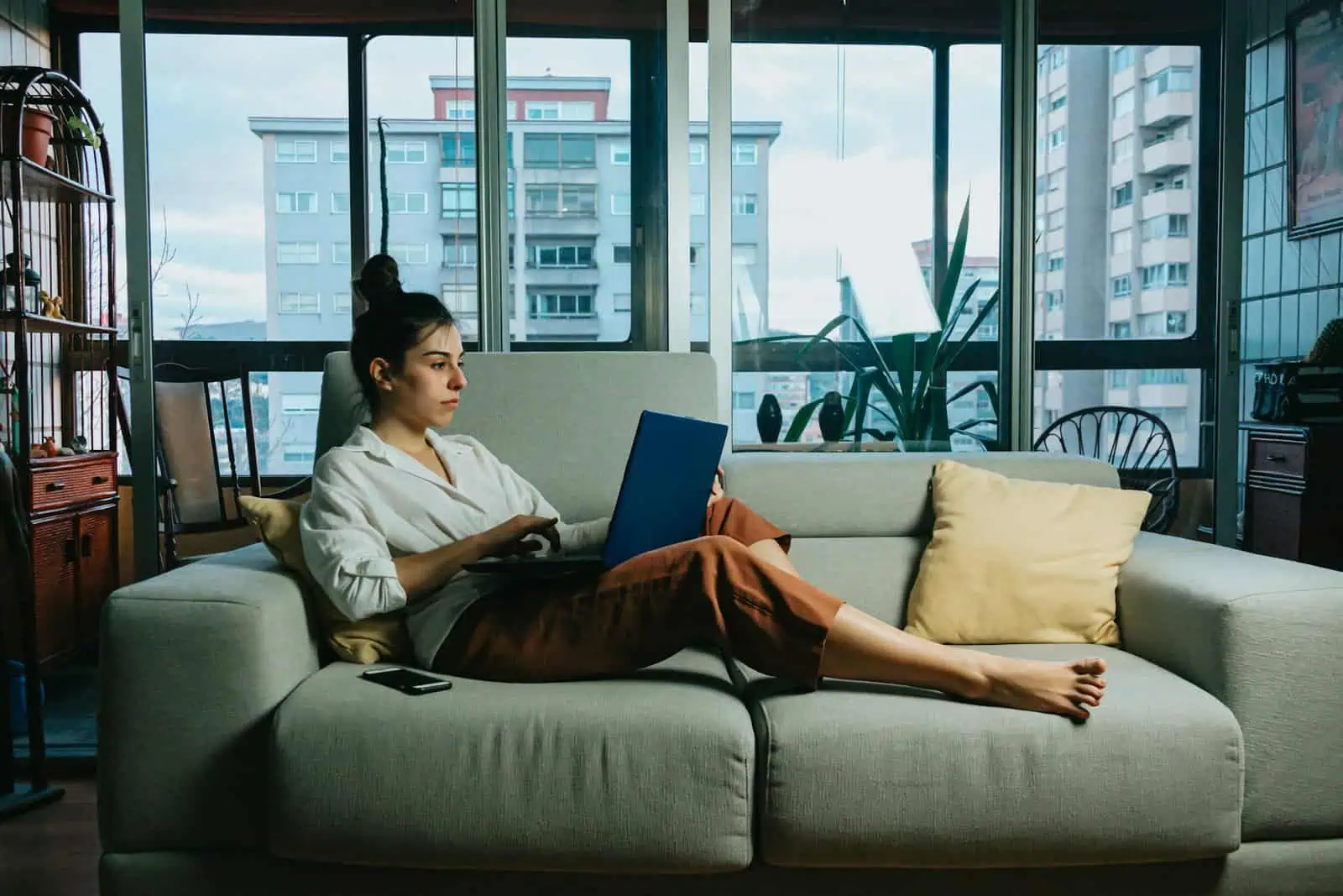 Young woman working on a laptop while relaxing on a sofa in a modern apartment, overlooking city buildings through large windows.