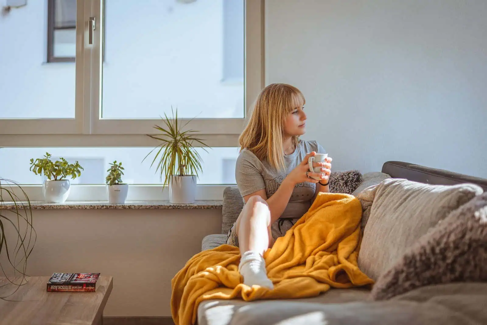 Relaxed girl enjoying a warm drink on sofa in living room with sunlight, houseplants, and cozy throw blanket, representing comfort, mental health, and well-being.