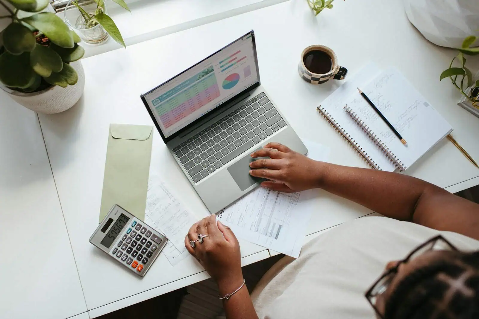 Financial planning and productivity tools on a white desk with a laptop, calculator, notebooks, and a cup of coffee, highlighting remote work, organisation, and financial management.