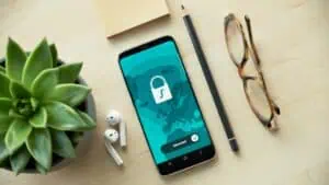 Smartphone displaying security app with a lock icon, surrounded by a plant, glasses, and stationery on a wooden desk, highlighting digital security and privacy.